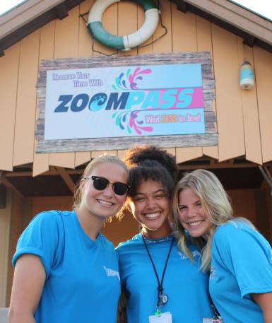 3 female team members stand in front of the ZoomPass sign smiling