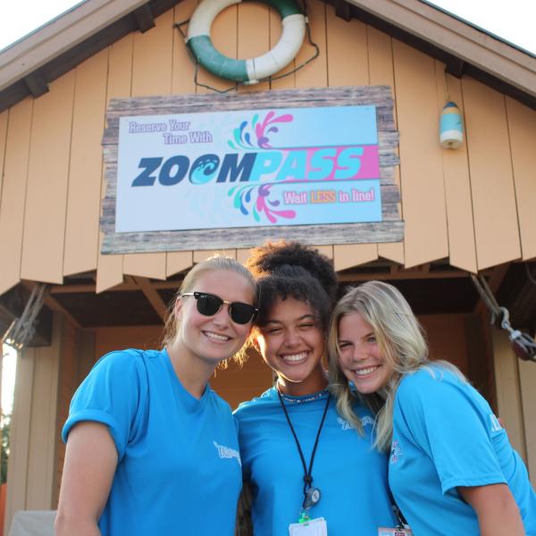 3 female team members stand in front of the ZoomPass sign smiling